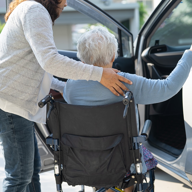 Une aide à domicile aide une personne âgée en fauteuil roulant à monter dans une voiture avec bienveillance.