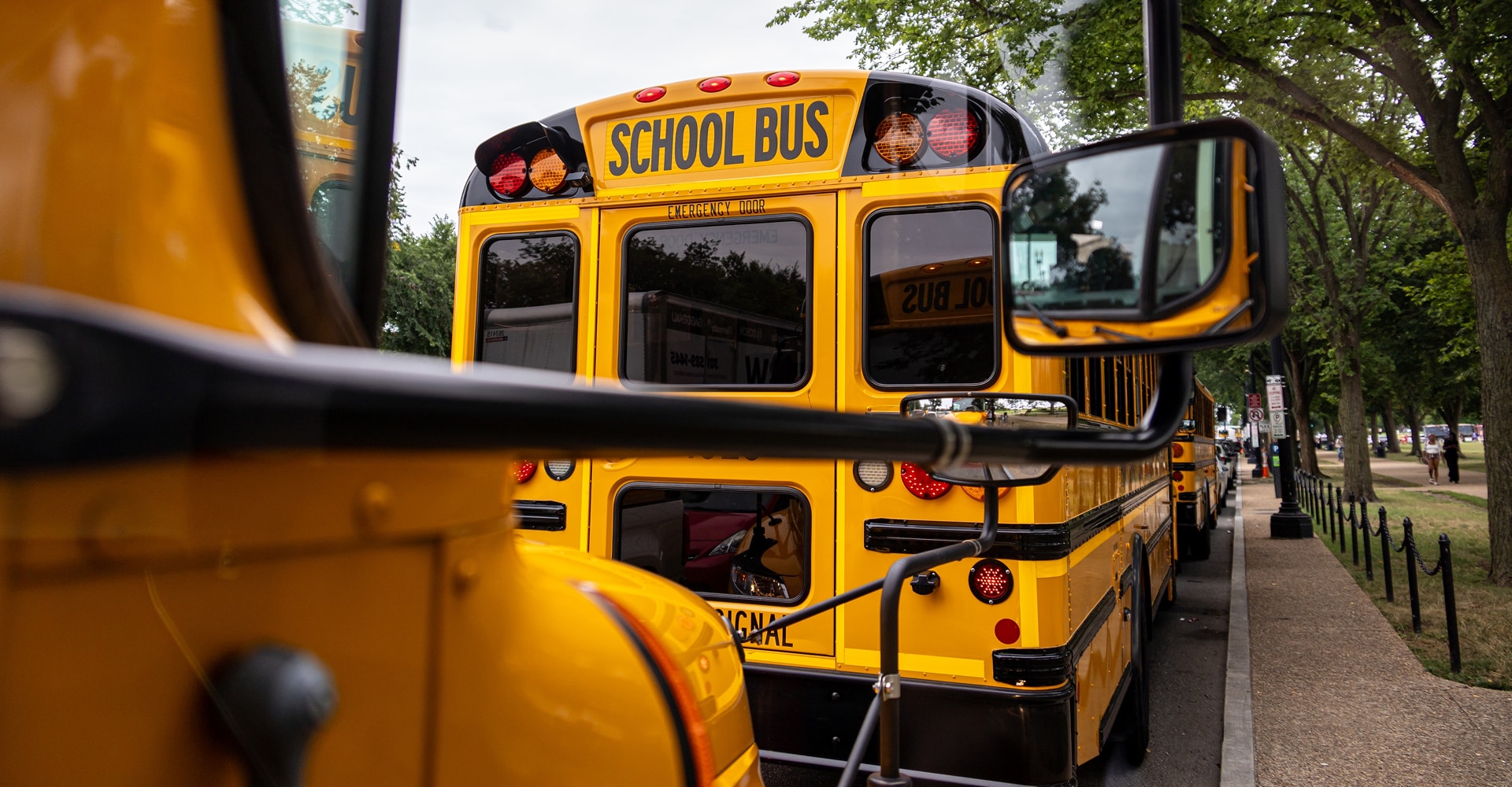 Arrière d'un bus scolaire jaune marqué SCHOOL BUS, encadré par le rétroviseur d'un autre véhicule en ville.