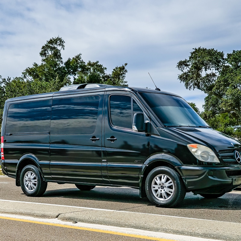 Van Mercedes Sprinter Noir de Luxe pour Transport VIP Van Mercedes Sprinter noir aux vitres teintées garé sur le bord d'une route sous un ciel nuageux.