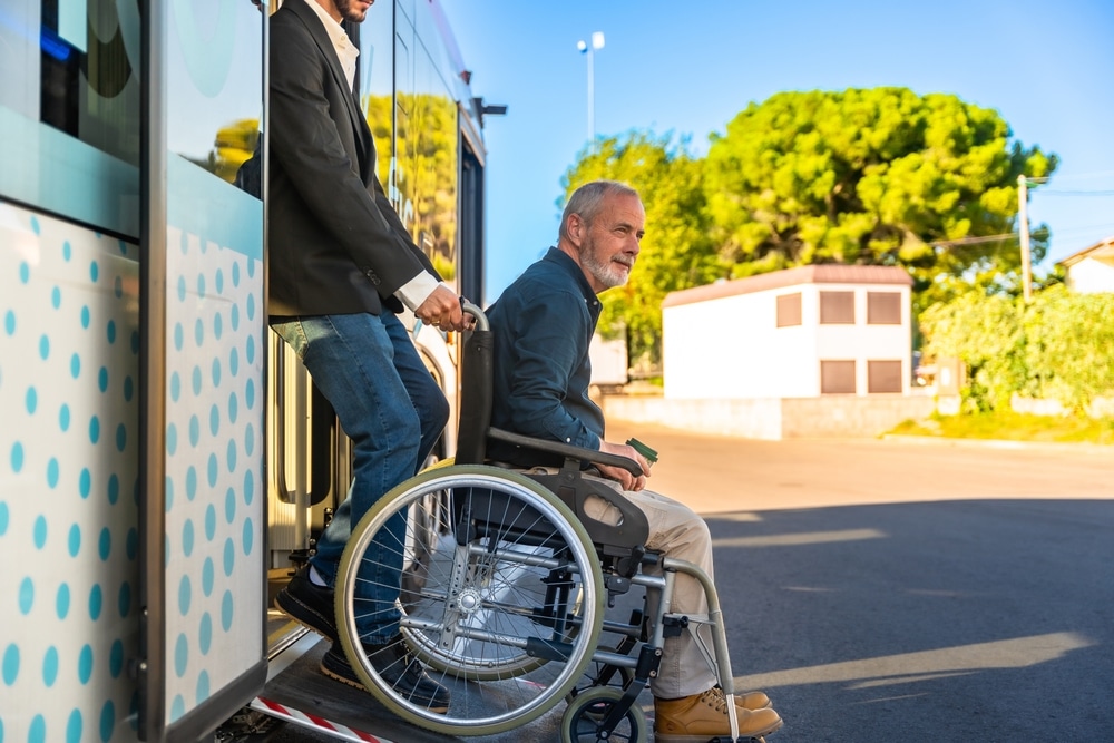 Homme aidant un senior en fauteuil roulant à descendre d'un bus via une rampe d'accès sous le soleil.