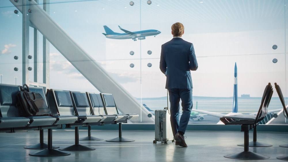Voyage d'affaires : homme attendant son vol à l'aéroport Un homme d'affaires avec sa valise regarde un avion décoller à travers la baie vitrée d'un aéroport.